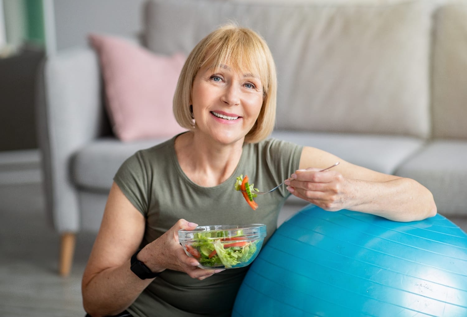 A middle-aged woman sitting on the floor, wearing activewear and eating a salad whilst resting against an exercise ball.
